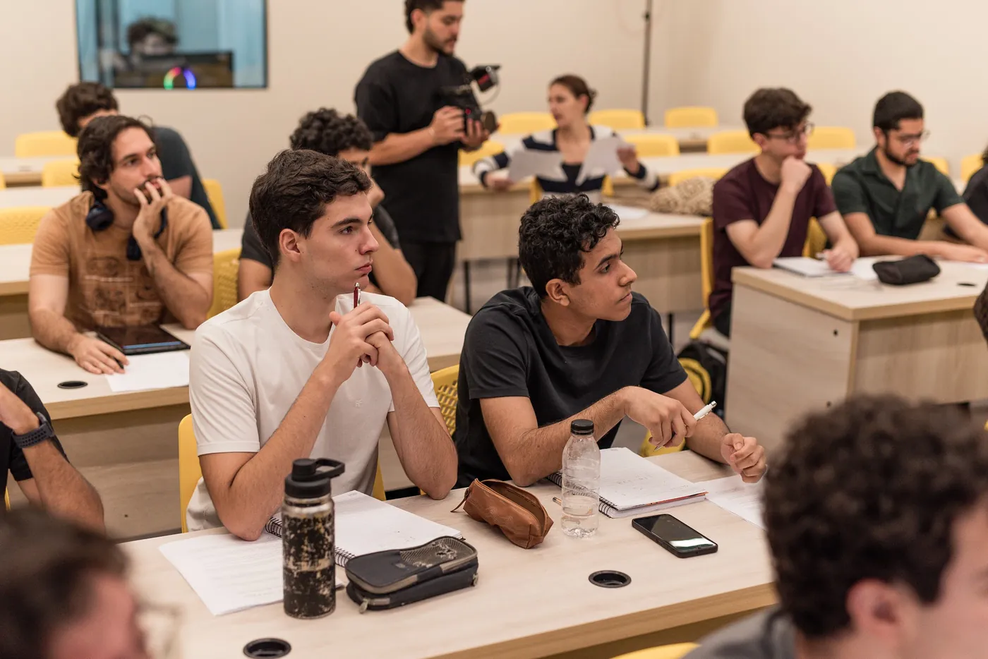 Alunos acompanhando a aula nas carteiras, com a câmera gravando ao fundo